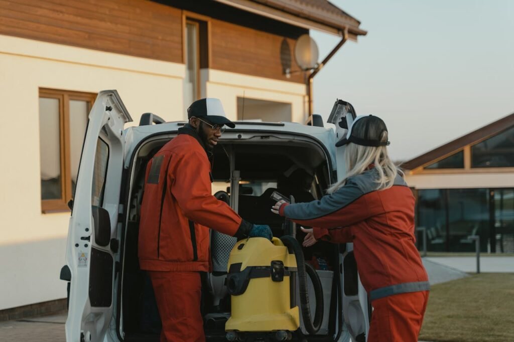 a-man-and-a-woman-getting-a-vacuum-cleaner-from-a-van-6195895 Two cleaners in red uniforms load a vacuum into a white van outdoors.