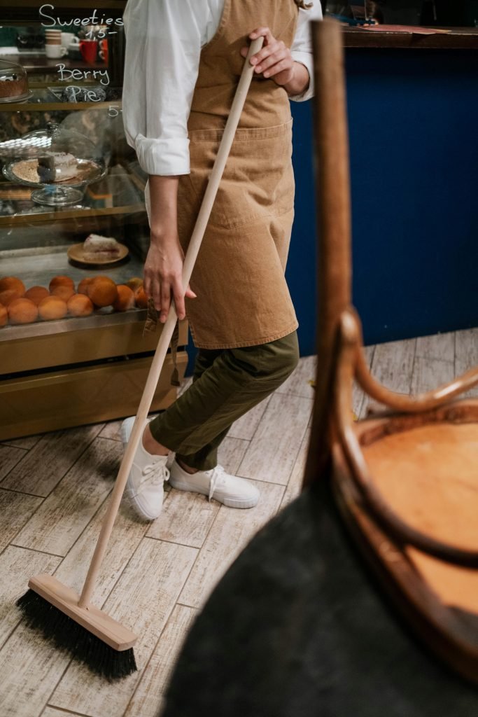 pexels photo 8441252 8441252 A person in an apron sweeps the wooden floor of a bakery with a broom, near pastries.